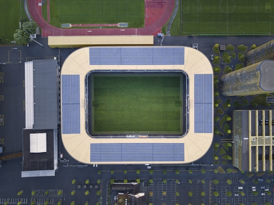An aerial view of a large stadium with a rectangular green field in the center, surrounded by solar panel installations on the roof. Adjacent to the stadium are parking lots, smaller buildings, and an athletic track. The surrounding area features orderly greenery and pathways.