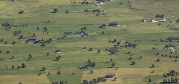 Aerial view of a rural landscape with expansive green fields and scattered houses. The terrain is flat with a few gently rolling areas, and patches of trees and small clusters of buildings dot the landscape. The fields are divided into irregular shapes, possibly indicating agricultural use.