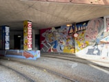 A vibrant mural under a bridge with colorful illustrations of musicians, musical instruments, and abstract patterns. The mural is painted on pillars and walls, featuring a headshot of a musician playing guitar and various musical notes. The setting includes railway tracks running through the area.
