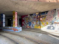 A vibrant mural under a bridge with colorful illustrations of musicians, musical instruments, and abstract patterns. The mural is painted on pillars and walls, featuring a headshot of a musician playing guitar and various musical notes. The setting includes railway tracks running through the area.