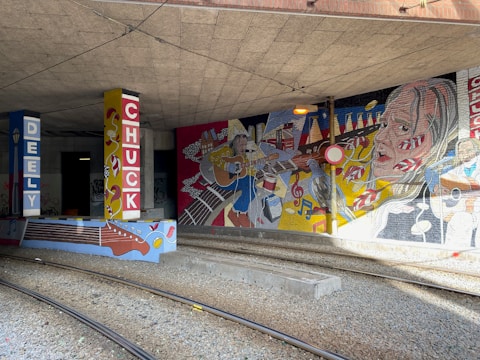 A vibrant mural under a bridge with colorful illustrations of musicians, musical instruments, and abstract patterns. The mural is painted on pillars and walls, featuring a headshot of a musician playing guitar and various musical notes. The setting includes railway tracks running through the area.