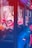 A row of claw machines filled with plush toys is bathed in vivid red and blue lighting. The machines are stocked with colorful stuffed animals, including characters wearing hats. The bright neon lights create a vibrant and slightly surreal atmosphere.