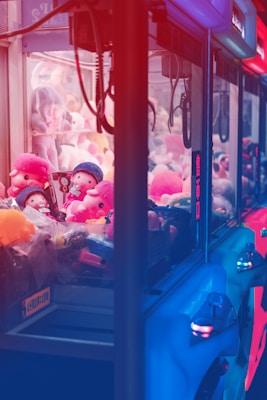 A row of claw machines filled with plush toys is bathed in vivid red and blue lighting. The machines are stocked with colorful stuffed animals, including characters wearing hats. The bright neon lights create a vibrant and slightly surreal atmosphere.