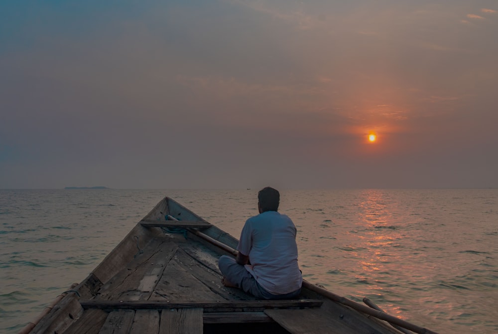 man in white shirt sitting on brown wooden boat on sea during sunset