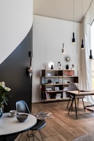 A modern interior space featuring a mix of minimalist and contemporary design elements. A round table with a marble top holds a teacup and bowl, accompanied by a dark chair. A wooden floor extends throughout the room. A tall shelf unit holds various decorative items, books, and personal mementoes. A desk with a chair is situated by a large window with sheer curtains, allowing natural light to flood the space. Pendant lights hang from the ceiling, and a geometric wall decoration adds interest.