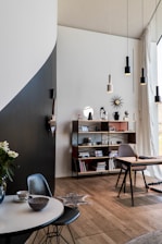 A modern interior space featuring a mix of minimalist and contemporary design elements. A round table with a marble top holds a teacup and bowl, accompanied by a dark chair. A wooden floor extends throughout the room. A tall shelf unit holds various decorative items, books, and personal mementoes. A desk with a chair is situated by a large window with sheer curtains, allowing natural light to flood the space. Pendant lights hang from the ceiling, and a geometric wall decoration adds interest.