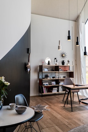 A modern interior space featuring a mix of minimalist and contemporary design elements. A round table with a marble top holds a teacup and bowl, accompanied by a dark chair. A wooden floor extends throughout the room. A tall shelf unit holds various decorative items, books, and personal mementoes. A desk with a chair is situated by a large window with sheer curtains, allowing natural light to flood the space. Pendant lights hang from the ceiling, and a geometric wall decoration adds interest.