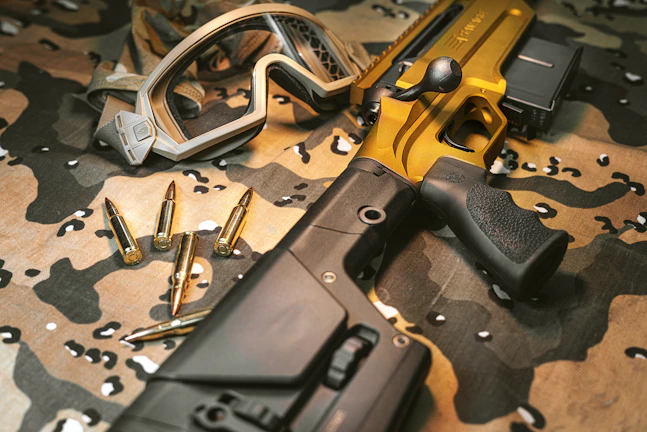 A tactical rifle with a metallic gold finish is placed on a camouflage fabric surface, accompanied by several bullets and a pair of protective goggles. The rifle features a robust black grip and adjustable stock.
