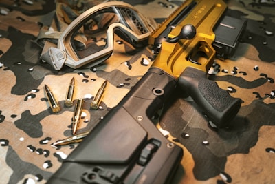 A tactical rifle with a metallic gold finish is placed on a camouflage fabric surface, accompanied by several bullets and a pair of protective goggles. The rifle features a robust black grip and adjustable stock.