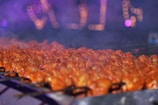 Skewers of marinated meat roasting over a grill with a smoky atmosphere. The background features blurred, colorful lights creating a festive ambiance.