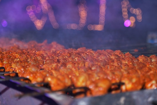 Skewers of marinated meat roasting over a grill with a smoky atmosphere. The background features blurred, colorful lights creating a festive ambiance.