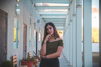 A young woman with long brown hair stands in a modern outdoor corridor. She is wearing an off-shoulder olive green top and black pants. Her expression is thoughtful, and she rests her chin on her hand. The corridor has a series of white columns and glass doors along one side, with wooden chairs and potted plants creating a serene environment.