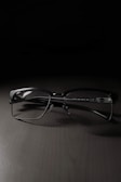 A pair of rectangular eyeglasses with thin black frames rests on a dark, smooth surface. The lighting creates a soft shadow beneath the frames, highlighting their sleek design.