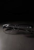 A pair of rectangular eyeglasses with thin black frames rests on a dark, smooth surface. The lighting creates a soft shadow beneath the frames, highlighting their sleek design.