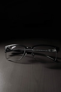 A pair of rectangular eyeglasses with thin black frames rests on a dark, smooth surface. The lighting creates a soft shadow beneath the frames, highlighting their sleek design.