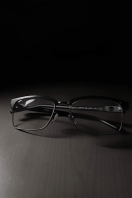 A pair of rectangular eyeglasses with thin black frames rests on a dark, smooth surface. The lighting creates a soft shadow beneath the frames, highlighting their sleek design.