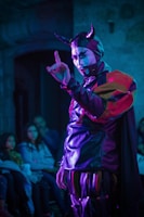 A person wearing a theatrical costume with horns and face paint is performing in front of a small audience. The costume consists of a shiny material with vibrant red, purple, and black colors. The setting is dimly lit, and the audience appears intrigued, seated in a small room.