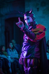 A person wearing a theatrical costume with horns and face paint is performing in front of a small audience. The costume consists of a shiny material with vibrant red, purple, and black colors. The setting is dimly lit, and the audience appears intrigued, seated in a small room.
