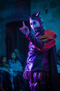 A person wearing a theatrical costume with horns and face paint is performing in front of a small audience. The costume consists of a shiny material with vibrant red, purple, and black colors. The setting is dimly lit, and the audience appears intrigued, seated in a small room.