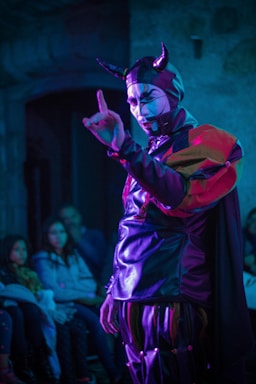 A person wearing a theatrical costume with horns and face paint is performing in front of a small audience. The costume consists of a shiny material with vibrant red, purple, and black colors. The setting is dimly lit, and the audience appears intrigued, seated in a small room.
