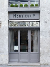 A stylish storefront with a gray painted facade and large glass doors. Above the doors, there is an awning and signage reading 'Monsieur P.' A row of neatly trimmed green shrubs in pots decorates the upper ledge.