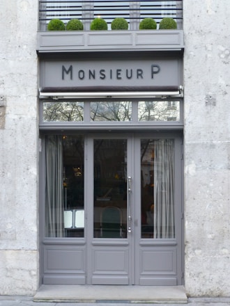 A stylish storefront with a gray painted facade and large glass doors. Above the doors, there is an awning and signage reading 'Monsieur P.' A row of neatly trimmed green shrubs in pots decorates the upper ledge.
