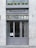 A stylish storefront with a gray painted facade and large glass doors. Above the doors, there is an awning and signage reading 'Monsieur P.' A row of neatly trimmed green shrubs in pots decorates the upper ledge.