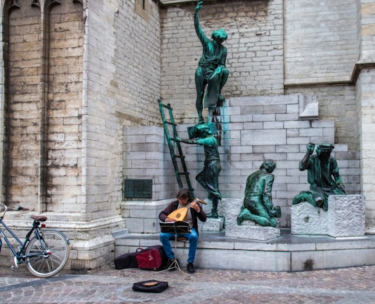 A musician sits on stone steps playing a stringed instrument. A bicycle is parked to the side, and behind the musician are bronze sculptures of people in various poses, set against an old stone wall.