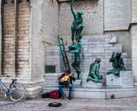 A musician sits on stone steps playing a stringed instrument. A bicycle is parked to the side, and behind the musician are bronze sculptures of people in various poses, set against an old stone wall.