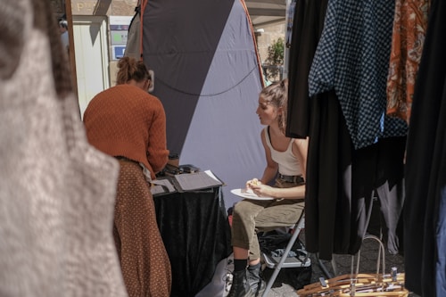 Two people are inside a stall or tent at an outdoor market. One person is sitting on a folding chair eating a pastry, while the other is standing, writing or organizing something on a table. There are clothes hanging around them, suggesting a market or fashion event.