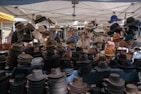 Under a canopy, a variety of hats are displayed on mannequin heads and tables at an outdoor market. The style of hats ranges from fedoras to wide-brimmed sun hats. Several people are browsing the selection, with one person looking at the hats thoughtfully. The scene is vibrant, with each hat offering different colors and textures.