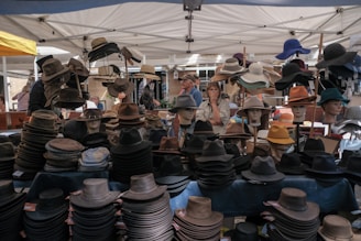 Under a canopy, a variety of hats are displayed on mannequin heads and tables at an outdoor market. The style of hats ranges from fedoras to wide-brimmed sun hats. Several people are browsing the selection, with one person looking at the hats thoughtfully. The scene is vibrant, with each hat offering different colors and textures.