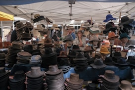 Under a canopy, a variety of hats are displayed on mannequin heads and tables at an outdoor market. The style of hats ranges from fedoras to wide-brimmed sun hats. Several people are browsing the selection, with one person looking at the hats thoughtfully. The scene is vibrant, with each hat offering different colors and textures.