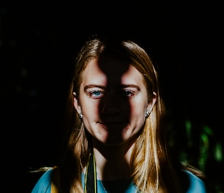 A person with long hair is standing against a dark background. A strong, narrow beam of light dramatically illuminates the center of their face, creating a striking contrast and sharp shadows.
