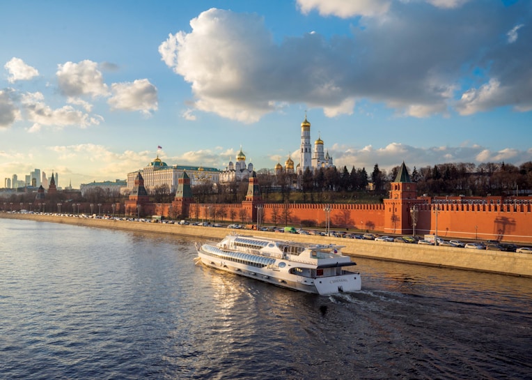 A large riverboat cruises along a wide river under a partly cloudy sky. On the riverbank, there is a long red brick wall with historic towers. In the background, a collection of elegant buildings with golden domes are visible, set against a backdrop of trees. The lighting suggests a late afternoon setting with the sun casting a warm glow.