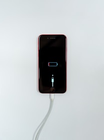 A smartphone with a red exterior is connected to a charging cable. The phone's screen displays a low battery icon with a charging symbol beneath it, indicating that the device is in the process of recharging.