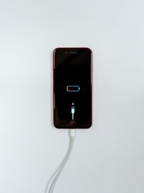 A smartphone with a red exterior is connected to a charging cable. The phone's screen displays a low battery icon with a charging symbol beneath it, indicating that the device is in the process of recharging.
