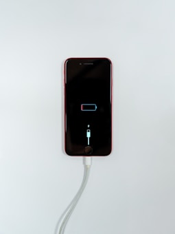 A smartphone with a red exterior is connected to a charging cable. The phone's screen displays a low battery icon with a charging symbol beneath it, indicating that the device is in the process of recharging.