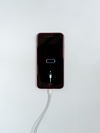 A smartphone with a red exterior is connected to a charging cable. The phone's screen displays a low battery icon with a charging symbol beneath it, indicating that the device is in the process of recharging.