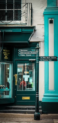 There is a storefront with a green door and window frames. The store has a sign in the window indicating it's open. Next to the door, there is another sign advertising the purchase of jewellery, silver, and gold. A decorative wrought iron balcony is visible above the store. In front of the store, there is a black signpost with pointers to several locations, including a racecourse, hospital, museum, and park. The building has accents of turquoise and green, and there is a sidewalk in front of the store.
