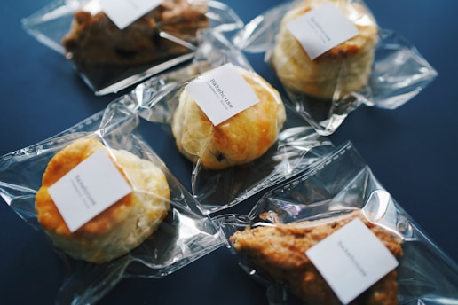 Several baked goods, such as pastries and scones, individually wrapped in clear plastic packaging with a white label on each, are arranged on a dark blue surface.