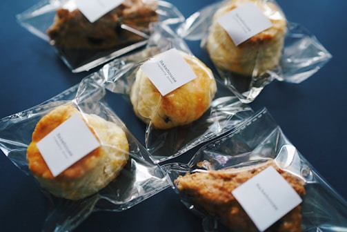 Several baked goods, such as pastries and scones, individually wrapped in clear plastic packaging with a white label on each, are arranged on a dark blue surface.