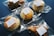 Several baked goods, such as pastries and scones, individually wrapped in clear plastic packaging with a white label on each, are arranged on a dark blue surface.