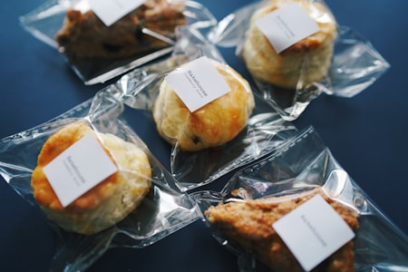 Several baked goods, such as pastries and scones, individually wrapped in clear plastic packaging with a white label on each, are arranged on a dark blue surface.