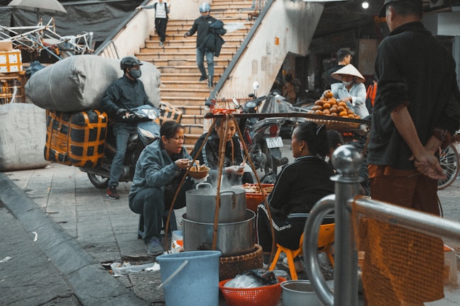 A bustling street scene with people gathered around a food vendor. The vendor's cart is laden with steaming pots and food items, with three people seated nearby eating. A person is making a purchase while others are passing by, and a scooter loaded with goods is parked on the left. The atmosphere is busy and lively.