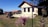 A rustic house with a red-tiled roof is situated in a rural setting with well-maintained grass and flower baskets in the foreground. There is an old machinery piece on the lawn, accompanied by scattered wooden logs. The sky is clear and blue, creating a serene atmosphere.