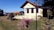 A rustic house with a red-tiled roof is situated in a rural setting with well-maintained grass and flower baskets in the foreground. There is an old machinery piece on the lawn, accompanied by scattered wooden logs. The sky is clear and blue, creating a serene atmosphere.