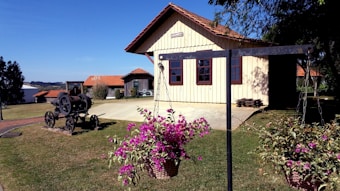 A rustic house with a red-tiled roof is situated in a rural setting with well-maintained grass and flower baskets in the foreground. There is an old machinery piece on the lawn, accompanied by scattered wooden logs. The sky is clear and blue, creating a serene atmosphere.