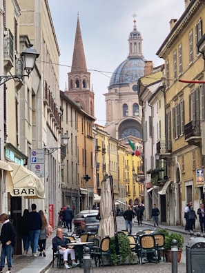 A picturesque street in an Italian town with historic architecture, featuring a prominent dome and a tall spire in the background. People are walking and sitting at an outdoor café with umbrellas, and there's an Italian flag hanging in the street. The buildings are adorned with balconies, lampposts, and various shop signs.