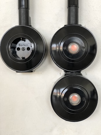 Three black circular electrical fixtures mounted on a light wall. The top-left fixture is a plug socket labeled 'Berker', while the other two have central buttons or lights.